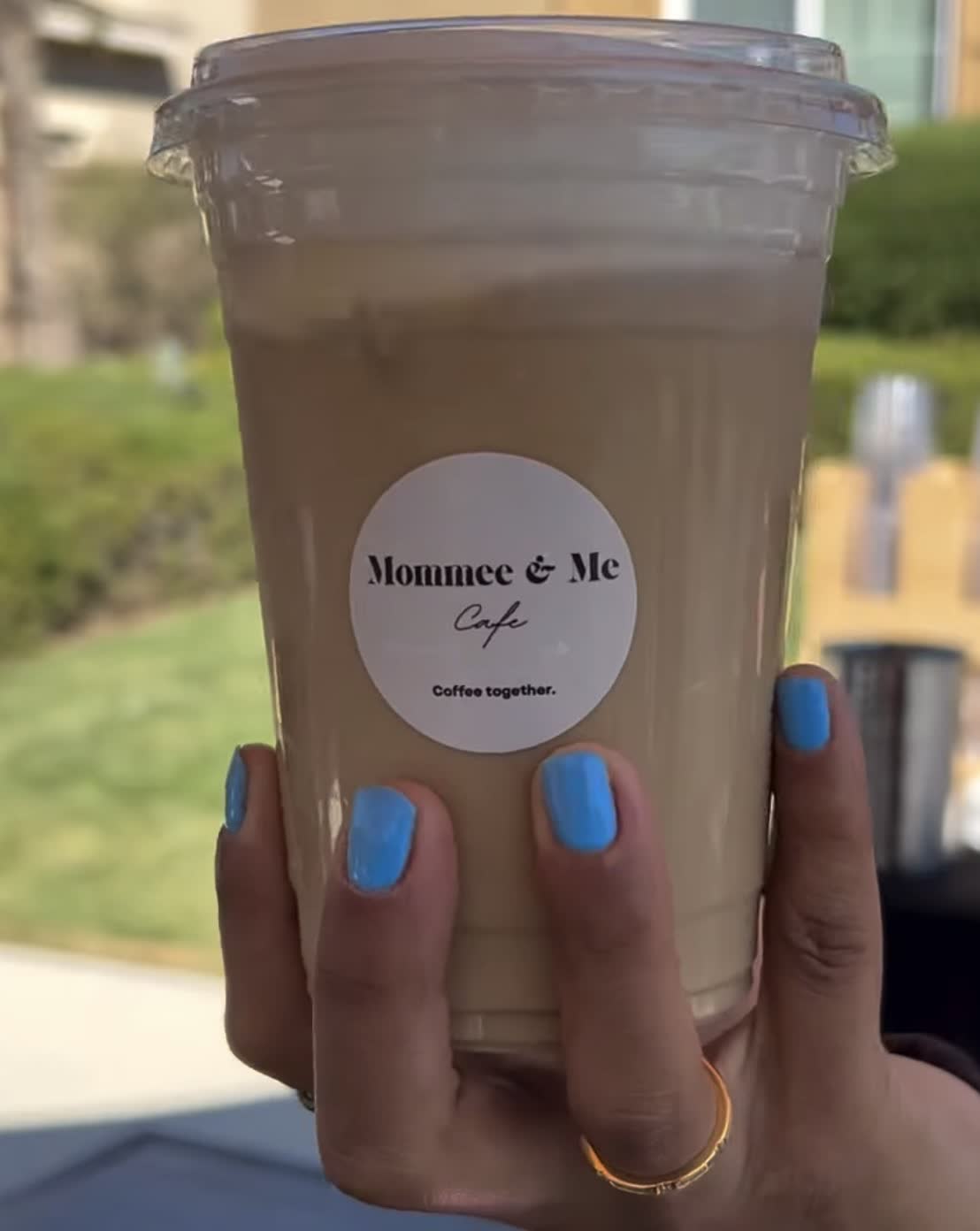 Iced latte with the round Mommee & Me Cafe sticker, held up at an outdoor event